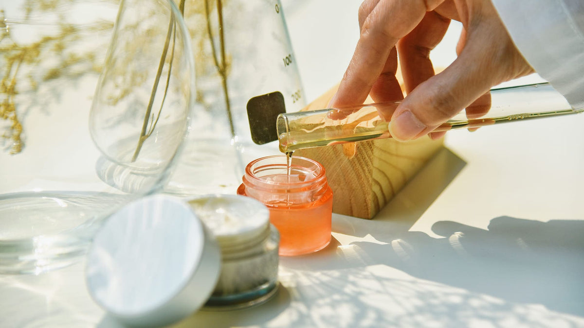 Person pouring liquid from a dropper into a small container on a light surface with flowers in the background