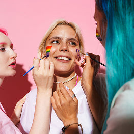 Two people applying makeup to a third person against a pink background